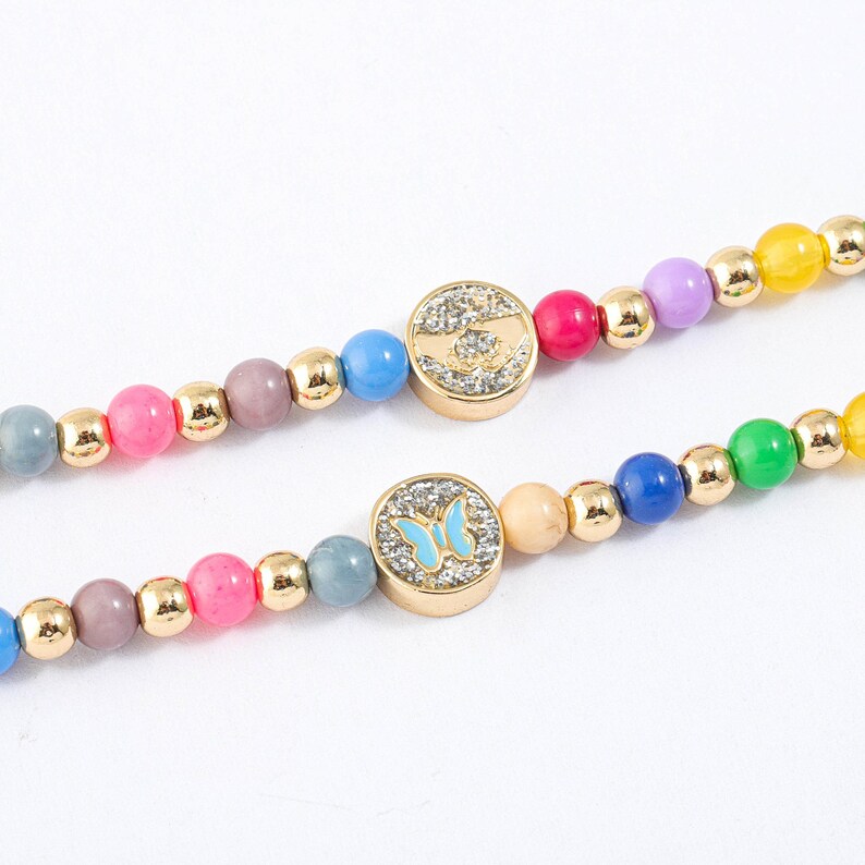 May include: Two colorful beaded bracelets with gold accents. One bracelet features a round charm with a handprint design, while the other bracelet features a round charm with a butterfly design.