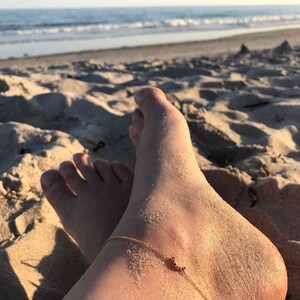May include: A person's bare feet and ankle with a gold anklet on a sandy beach. The anklet has a small crescent moon charm.