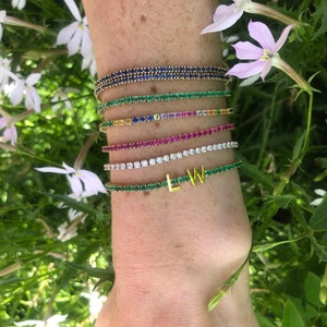 Puede incluir: Una muñeca de mujer con una pila de pulseras de oro con piedras preciosas de diferentes colores. Las pulseras son una doble fila de zafiros azules, una fila simple de esmeraldas verdes, una fila simple de zafiros amarillos, una fila simple de zafiros rosas, una fila simple de rubíes rojos, una fila simple de diamantes transparentes y una fila simple de esmeraldas verdes con letras doradas "LW" en el centro.