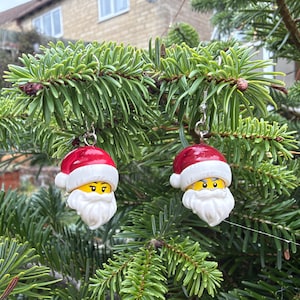 May include: A pair of earrings featuring miniature Santa Claus heads with yellow faces, white beards, and red hats. Each head is designed with a classic toy figure aesthetic, complete with black eyes and eyebrows, suspended from silver-toned metal hooks.