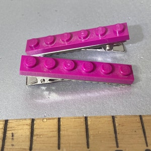 May include: Two purple plastic hair clips with a row of five Lego bricks attached to each clip.