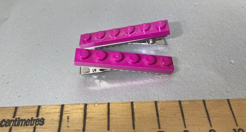 May include: Two purple plastic hair clips with a row of five Lego bricks attached to each clip.
