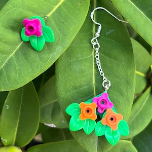 May include: A pair of floral earrings featuring a bright pink and orange flower design with green leaves. The earrings are set against a backdrop of green leaves, creating a vibrant and natural aesthetic. The earrings are a unique accessory.