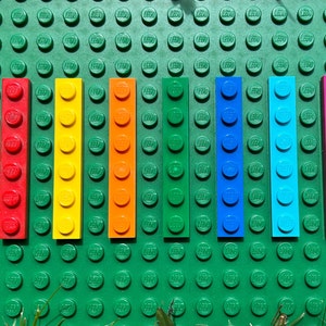 May include: A row of eight colorful Lego bricks, arranged in a rainbow pattern from red to pink. The bricks are all the same size and shape, and they are set against a green background.