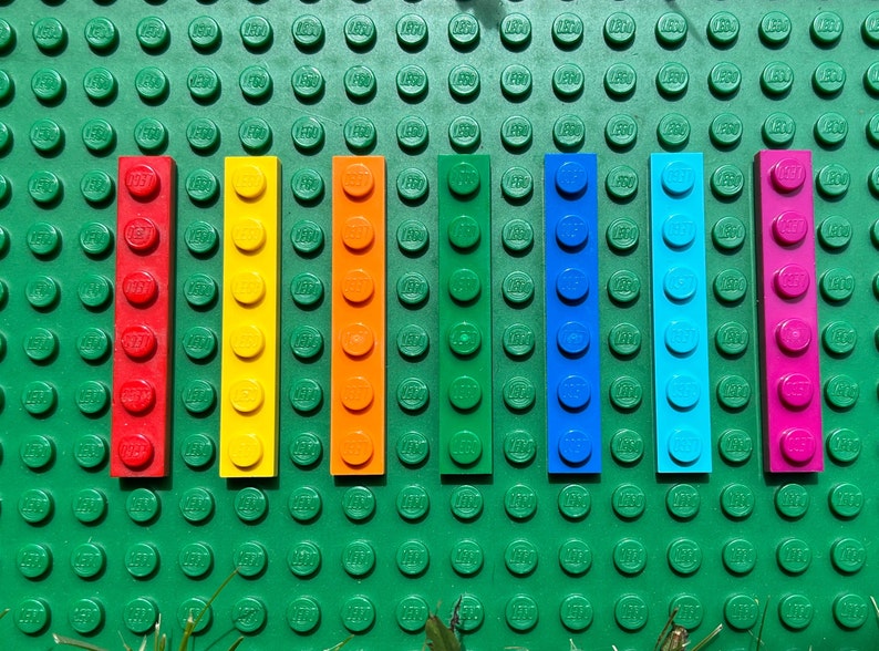 May include: A row of eight colorful Lego bricks, arranged in a rainbow pattern from red to pink. The bricks are all the same size and shape, and they are set against a green background.