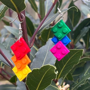 May include: A pair of handmade earrings featuring colorful Lego bricks. One earring has red, orange, and yellow bricks, while the other has green, blue, and pink bricks. The earrings hang from silver-colored hooks.