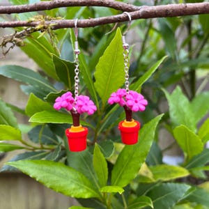 May include: A pair of dangle earrings featuring miniature pink flower plants in red pots. The earrings hang from silver-colored hooks and chains. The background is a natural setting with green leaves and a brown branch.