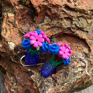 Könnte beinhalten: Ein Paar handgefertigte Ohrringe mit Miniatur-Blumenarrangements. Jeder Ohrring hat einen blauen Topf mit einer rosa Blume und blauen Akzenten. Die Ohrringe bestehen aus kleinen Kunststoffteilen und haben silberfarbene Haken.