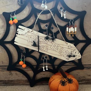 May include: A black spiderweb-shaped earring display rack holding various Halloween-themed earrings, including pumpkins, skulls, bones, spiders, and candles. A wooden sign with the text "TRICK OR TREAT" hangs in the center with a small orange pumpkin below.
