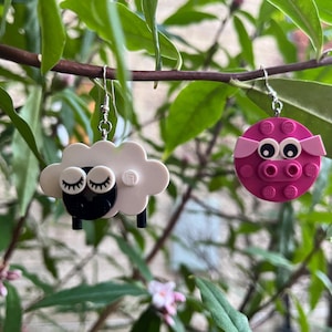 May include: A pair of whimsical earrings. One earring is a white sheep with black accents and closed eyes. The other is a pink pig with large eyes and ears. Both earrings are suspended from silver hooks, set against a backdrop of green leaves.