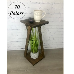 May include: A dark brown wooden side table with a square top and a geometric base. A white mug, a remote control, and a small potted plant are on the table. The text "10 Colors" is in a circle.