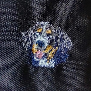 May include: Embroidered patch of a dog's face, using blue, grey, yellow, and pink thread on navy fabric. The dog's face is intricately stitched, highlighting its features with detailed embroidery.