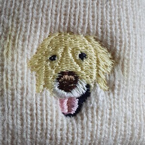 Puede incluir: Gorro de punto color crema con un bordado de la cara de un perro golden retriever. La cara del perro está detallada con ojos negros, una nariz marrón y una lengua rosa. El gorro está hecho de un material suave y texturizado.