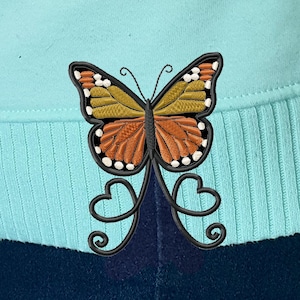 May include: A light blue sweater with a black embroidered butterfly design. The butterfly has orange, brown, and yellow wings.
