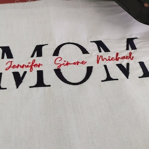 Może przedstawiać: Biały materiał z czarnymi haftowanymi literami tworzącymi napis "MOM" z nazwami "Jennifer", "Simone" i "Michael" napisanymi na czerwono wewnątrz liter.