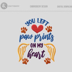 以下が含まれることがあります： 青い、紫色の、そしてオレンジ色の「You left paw prints on my heart」というテキストが刺繍されたデザイン。赤いハートがデザインの中心にあり、ハートの両側にオレンジ色の肉球の跡が2つあります。ハートとテキストの後ろには、黄色の羽が2枚あります。
