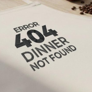 May include: Off-white apron with the text "ERROR 404 DINNER NOT FOUND" in black. A wooden spoon, a vintage stopwatch, and coffee beans are on the wooden surface. The apron has a neck strap and waist ties.