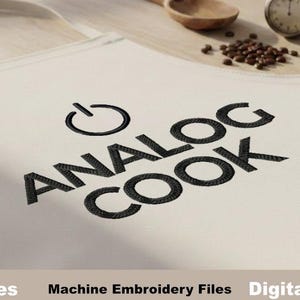 May include: Off-white apron with the words "ANALOG COOK" embroidered in black. The apron also features a power symbol above the text. A wooden spoon, a vintage stopwatch, and coffee beans are on the wooden surface.