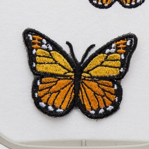 Pode incluir: Dois patches de borboleta bordados em um aro de bordado branco. As borboletas são amarelas e laranja com contornos pretos.