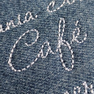 Może przedstawiać: Zbliżenie niebieskiego dżinsu z białym haftowanym tekstem. Słowo "Café" jest wyraźnie widoczne, z częściowo widocznym innym tekstem kursywą. Szycie jest szczegółowe i tworzy efekt tekstury.