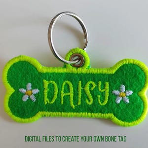 Könnte beinhalten: Grünes, knochenförmiges Hundehalsband mit einem lindgrünen Rand, auf dem der Name "Daisy" in lindgrün mit weißen Gänseblümchen auf beiden Seiten gestickt ist.
