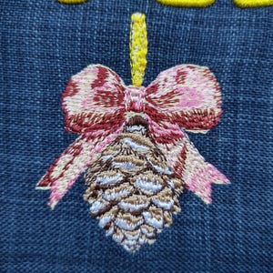 May include: Embroidered pinecone design with a pink and burgundy bow, and a yellow stem, on a blue denim background. The pinecone is detailed with shades of brown and white. The embroidery is likely for a decorative item or patch.