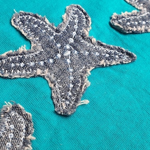 May include: Close-up of a teal fabric with three denim starfish appliqués. The starfish are stitched with white thread and have a frayed edge. The fabric has a textured appearance.