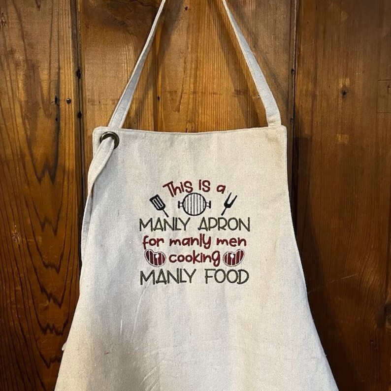 May include: A white linen apron with the text "This is a MANLY APRON for manly men cooking MANLY FOOD" embroidered in brown and red thread. The apron has a brown loop at the top and is hanging on a wooden surface.