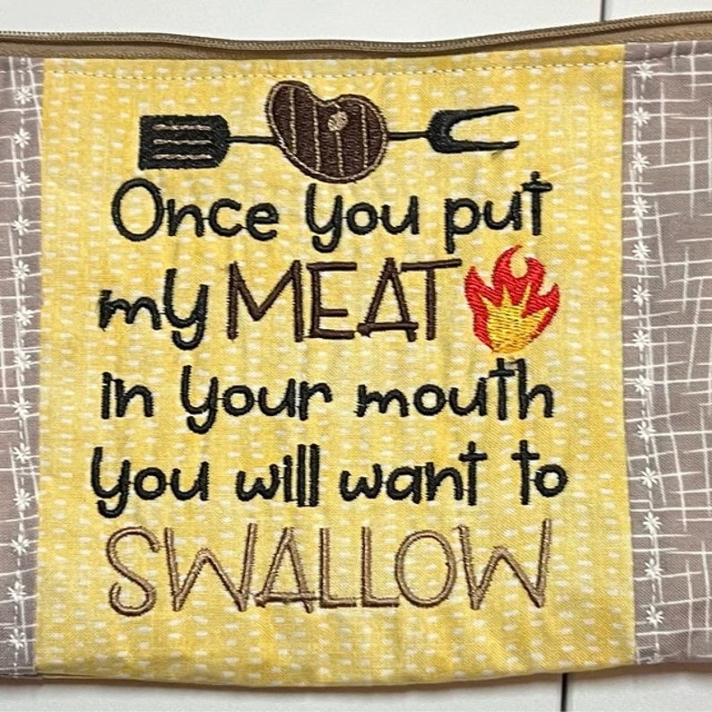 May include: A yellow fabric zipper pouch with a brown and white checkered border. The pouch has a white embroidered design with the text "Once you put my MEAT in your mouth you will want to SWALLOW". The design also includes a red and yellow flame and a black silhouette of a spatula, a steak, and a fork.