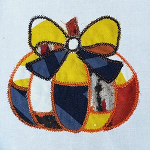May include: A colorful, patchwork pumpkin design with a yellow bow and orange stitching. The pumpkin is made of various fabric pieces in yellow, blue, red, and white. The design is on a white background.
