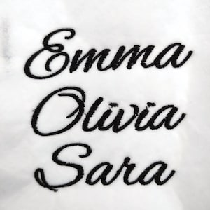 Puede incluir: Tela blanca con nombres bordados en negro: Emma, Olivia y Sara. El texto "real embroidery test" está encima de los nombres. El bordado está en una fuente cursiva, mostrando una muestra de texto personalizado.