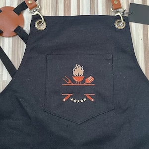 May include: Black canvas apron with a large pocket featuring an embroidered design of a grill, flames, and grilling tools. The design also includes five stars.
