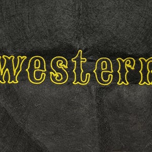 May include: Black fabric with the word "western" embroidered in yellow. The text has a decorative, vintage-style font. The fabric has a textured appearance.