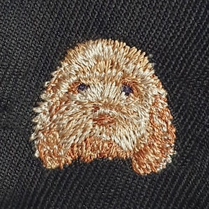 May include: Close-up of an embroidered dog face with a fluffy, light brown and tan design. The dog's eyes are small and blue, and the embroidery is on a dark grey fabric.