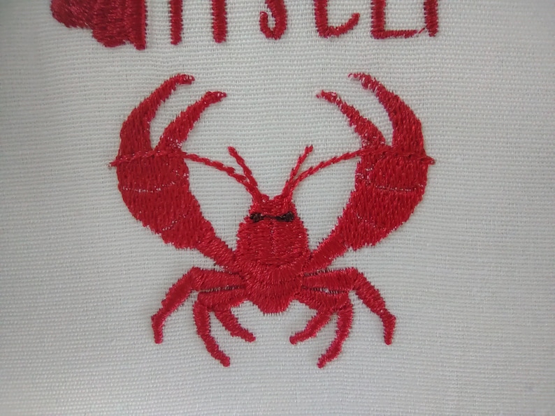 Pode incluir: Design de lagosta bordada em vermelho sobre um fundo de tecido branco.