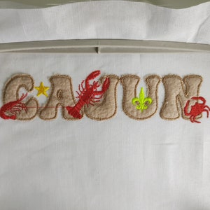 Pode incluir: Desenho bordado da palavra "CAJUN" em castanho com acentos vermelhos e amarelos. O desenho inclui uma estrela, um lagostim, uma flor-de-lis e um caranguejo.