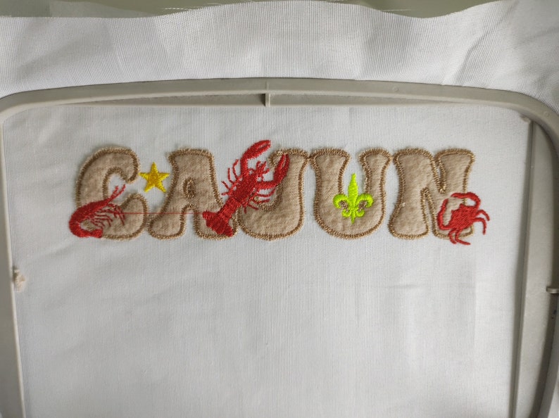 Pode incluir: Desenho bordado da palavra "CAJUN" em castanho com acentos vermelhos e amarelos. O desenho inclui uma estrela, um lagostim, uma flor-de-lis e um caranguejo.