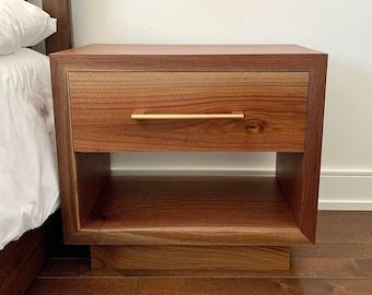 May include: A wooden nightstand with a single drawer and a lower shelf. The drawer has a gold metal handle. The nightstand is made of dark wood with a natural wood grain.