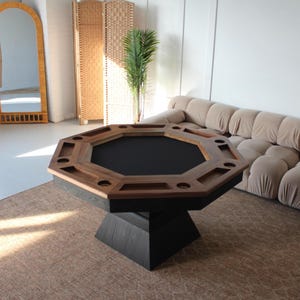 Puede incluir: Una mesa de póquer octogonal con una superficie de juego negra y un borde de madera oscura. La mesa tiene portavasos incorporados y una base de pedestal negra. La mesa está sobre una alfombra de color neutro.