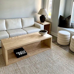 Puede incluir: Una mesa de centro de madera clara con un estante inferior y un libro negro con el texto "TOM FORD" en la parte superior. La mesa está en una sala de estar con un sofá blanco, un otomano blanco y una alfombra.