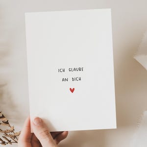 Op de afbeelding: Een witte kaart met de Duitse tekst "ICH GLAUBE AN DICH" en een klein rood hart. De kaart wordt door een hand vastgehouden, met gedroogde varen en wit lint op de achtergrond.