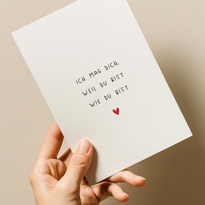 May include: A white card with black text and a small red heart is held in a hand. The text reads "ICH MAG DICH, WEIL DU BIST, WIE DU BIST."