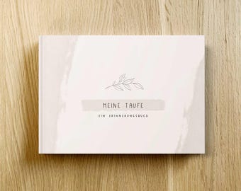 May include: A hardcover book with the text "MEINE TAUFE" and "EIN ERINNERUNGSBUCH" in a minimalist design. The cover is off-white with a light beige watercolor wash and a small leaf illustration. The book is on a wooden surface.