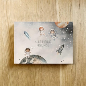 Könnte beinhalten: Ein Kinderbuch mit Weltraumthema. Das Cover zeigt Illustrationen von Astronauten, Planeten, einer Rakete und einem Sternschnuppen vor einem Sternenhintergrund. Der Text "ALLE MEINE FREUNDE" ist auf dem Cover abgebildet.
