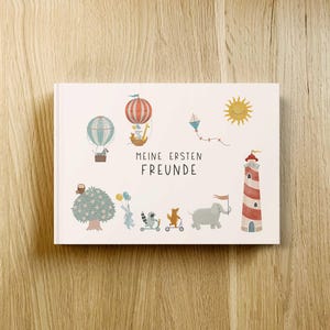 Könnte beinhalten: Ein weißes Hardcover-Buch mit dem deutschen Text "Meine ersten Freunde" und Illustrationen eines Heißluftballons, Tieren, eines Leuchtturms und einer Sonne. Das Buch liegt auf einer Holzoberfläche.