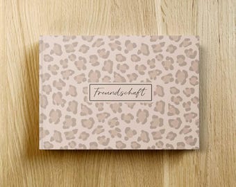 May include: A closed book with a light pink cover featuring a leopard print pattern in shades of beige and taupe. The word "Freundschaft" is printed in a black, rectangular frame on the cover. The book is set against a wooden background.