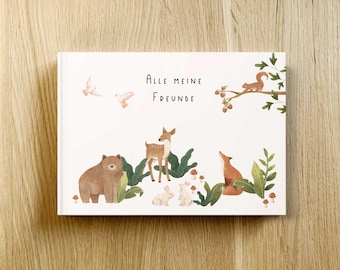 May include: A hardcover book with the title "ALLE MEINE FREUNDE" and illustrations of woodland animals. The cover features a brown bear, deer, fox, rabbits, and a squirrel on a branch, with birds in the sky. The book is on a wooden surface.