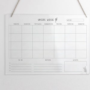 May include: A white acrylic weekly planner with black lines and text. The text says "UNSERE WOCHE" at the top and includes the days of the week in German: Montag, Dienstag, Mittwoch, Donnerstag, Freitag, Samstag, Sonntag. There are sections for "TO DO", "NOTIZEN", and "WICHTIG".