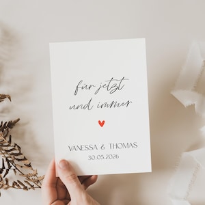 Può includere: Un biglietto color crema con il testo "für jetzt und immer" e un cuore rosso. I nomi "VANESSA & THOMAS" e la data "30.05.2026" sono stampati sotto. Una mano tiene il biglietto, con foglie secche e nastro bianco sullo sfondo.