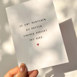 May include: A white card with black text and a small red heart. The text reads, "ES GIBT MENSCHEN, DIE PASSEN EINFACH PERFEKT INS HERZ." The card is held by a hand, with sunlight and shadows in the background.